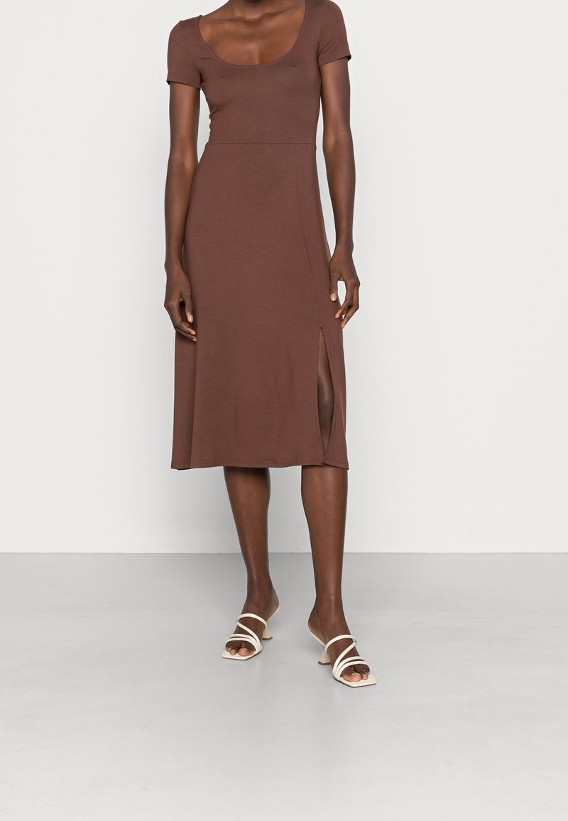 Kvinna i en brun midiklänning med korta ärmar och en sidoklyvning, i kombination med vita sandaler med låg klack och remmar, står på ett ensamt golv.