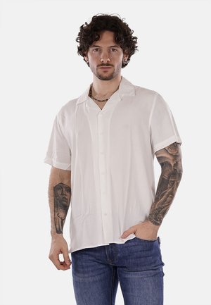 Camicia bianca a maniche corte con bottoni davanti, colletto e texture liscia. Indossata con jeans blu in denim, il modello presenta tatuaggi sulle braccia.