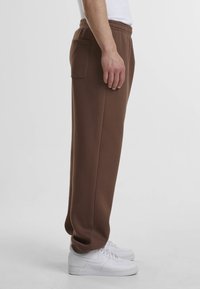Pantalons de survêtement marron en tissu doux, avec une poche latérale et une taille élastique, associés à des baskets blanches. Texture lisse.