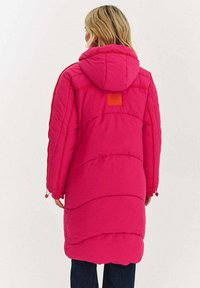 Dlouhý růžový péřový kabát s kapucí, s prošitými částmi a čtvercovou nášivkou na zádech. Nastavitelné šňůrky na spodním okraji.