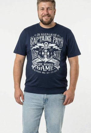 Uśmiechnięty mężczyzna z brodą, ubrany w granatowy t-shirt z białym wzorem kotwicy i tekstem o tematyce morskiej, w połączeniu z jasnoniebieskimi jeansami.