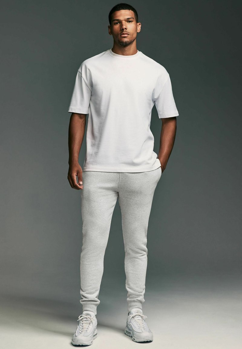 Biely bavlnený tričko s krátkymi rukávmi v kombinácii so svetlo šedými joggermi z mäkkej tkaniny. Biely tenisky dotvárajú celý outfit.