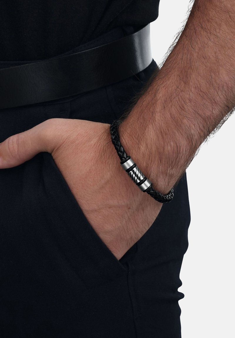 La mano de un hombre lleva una pulsera de cuero trenzado negro con un broche metálico, descansando en el bolsillo de unos pantalones negros con un cinturón negro.
