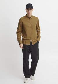 Cămașă maro cu nasturi, fabricată dintr-un material moale, asortată cu pantaloni negri și adidași albi. Modelul poartă o șapcă neagră.