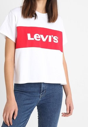 Personne portant un T-shirt blanc court avec un rectangle rouge et le logo blanc Levi's sur la poitrine, associé à un jean bleu.