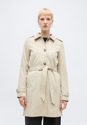 Blondynka w beżowym płaszczu trencz z paskiem, guzikami i detalami na pasku, stojąca na jasnym, jednolitym tle.