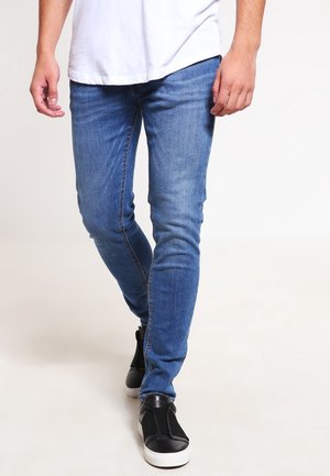 Mann, der in schlanken blauen Jeans, weißem T-Shirt und schwarzen Slip-On-Sneakern mit weißen Sohlen auf hellem, schlichten Hintergrund geht.