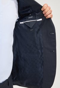 Czarna marynarka z wzorzystym granatowym wnętrzem, z etykietą slim fit i widocznymi detalami szwów; trzymana przez osobę, ukazująca podszewkę.