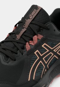 Zwarte sportieve schoen met een gestructureerde bovenkant, voorzien van roze en koperkleurige accenten en dunne zwarte veters. Kenmerkend logo en grafische elementen zichtbaar.