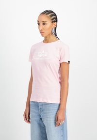 Světle růžové bavlněné tričko s bílým logem "Alpha Industries", krátké rukávy a volný střih. Vytvořeno s světle modrým denimem.