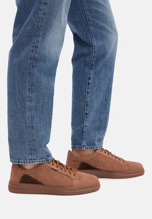 Sneakers in suede marrone con suole in gomma marrone, caratterizzate da un pannello laterale in un marrone scuro a contrasto e lacci marroni. Indossati con jeans blu.