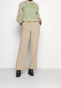 Zelená žebrovaná vesta přes květinovou blůzu, kombinovaná s béžovými širokými kalhotami s pruhy a černými botami na podpatku. Texturovaná látka a vrstvený design.