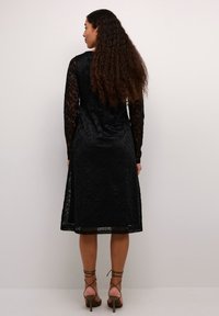 Black lace dress with long, sheer sleeves and floral texture, knee-length, featuring a flared skirt and a back slit, paired with tan heels.