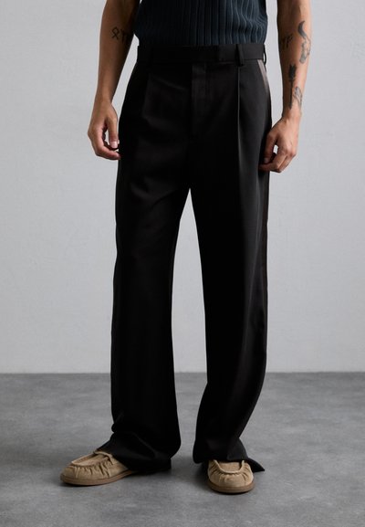 Sorte plisserede bukser med en skræddertilpasset pasform, lavet af glat stof; båret med beige slip-on sko med samlet detaljer.