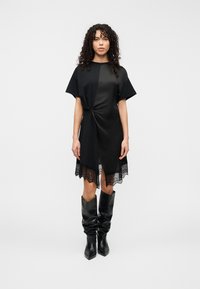 Μαύρο φόρεμα με κόμπο στο μπροστινό μέρος, κοντά μανίκια και δαντέλα στο κάτω μέρος. Συνδυάζεται με μαύρες, χαλαρές μπότες μέχρι το γόνατο.