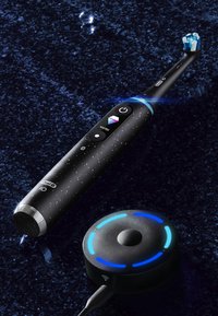 Brosse à dents électrique avec un corps noir tacheté, des accents bleus et des boutons de contrôle. Comprend une base de charge ronde avec des anneaux lumineux.