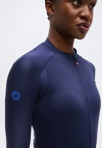 Κοντινό πλάνο ενός ατόμου που φοράει εφαρμοστό μπλε σκούρο μακρυμάνικο πουκάμισο με φερμουάρ στο μπροστινό μέρος και μικρό στρογγυλό λογότυπο σκορπιού στον πάνω βραχίονα.