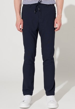 Homme portant un pantalon slim bleu marine avec taille à cordon de serrage et des baskets blanches, debout devant un fond blanc.