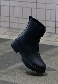 Black ankle boot made of smooth leather with a chunky rubber outsole, featuring a visible zipper and a rounded toe design.