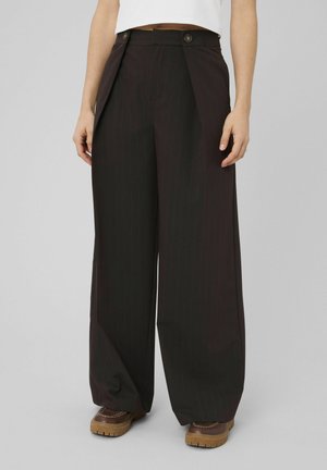 Femme portant un pantalon noir à larges jambes à fines rayures avec des boutons au niveau de la taille, associé à des chaussures marron sur un fond gris.
