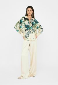 Blumenbluse mit blauen und grünen Druckmotiven, cremefarbener Grundfarbe, oversized Ärmeln, kombiniert mit cremefarbenen weiten Hosen; das Model steht mit den Händen in den Hüften.