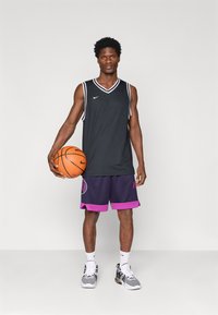 Μαύρη αμάνικη φανέλα μπάσκετ με λευκές λεπτομέρειες, συνδυασμένη με μωβ σορτς που έχουν ροζ περιθώριο και λογότυπο, κρατώντας μια πορτοκαλί μπάλα μπάσκετ.