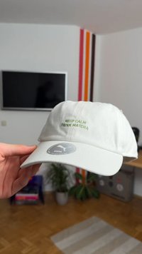 Gaiši pelēka kokvilnas cepure ar izliekto maliņu. Priekšpusē ir zaļā krāsā izšīts uzraksts "SAGLABĀ RAUPSMU, DZERS MĀČU".