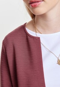 Zbliżenie na osobę ubraną w żeberkowaną mauve kurtkę, białą koszulkę i złoty naszyjnik z małym wisiorkiem.