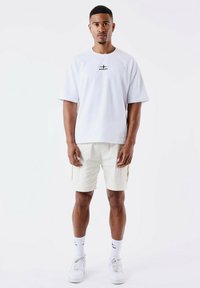 Camiseta blanca de gran tamaño con un pequeño logo negro, combinada con pantalones cortos de cargador beige claro que cuentan con bolsillos laterales y costuras en contraste.