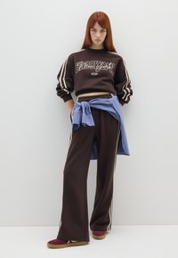 Sudadera marrón con estampado de "Brooklyn", pantalones de chándal marrones y una camisa a cuadros azul atada a la cintura. Zapatos rojos con suelas marrones.