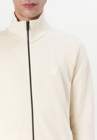 Felpa beige con zip e colletto alto. Presenta un logo discreto sul petto, una zip nera e un tessuto dalla texture liscia senza motivi visibili.