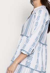 Lekka, bawełniana sukienka w kolorze białym w niebieskie pionowe paski. Ozdobiona geometrycznymi wzorami i frędzlami na rękawach oraz u dołu.