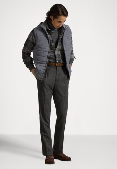 Grå quiltet vest over en ternet skjorte, kombineret med mørke skræddersyede bukser og brune loafers, sat mod en ensfarvet hvid baggrund.
