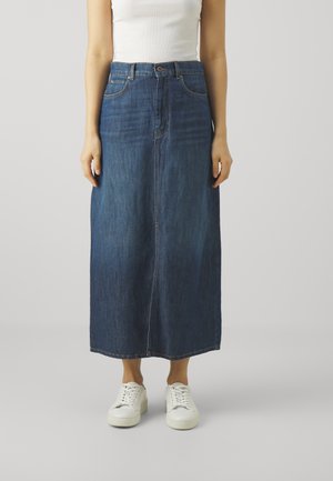 Femme portant un haut blanc sans manches, une longue jupe en jean bleu foncé et des baskets blanches, debout devant un fond uni.