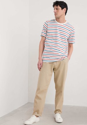 Maglietta a righe con linee orizzontali rosse, blu e bianche, colletto a girocollo, abbinata a pantaloni beige e sneakers bianche.