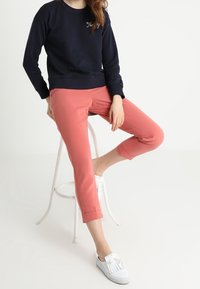 Sudadera de marina con texto bordado, pantalones cortos de coral, y zapatillas blancas con acentos de borlas. Modelo sentado en un taburete blanco.