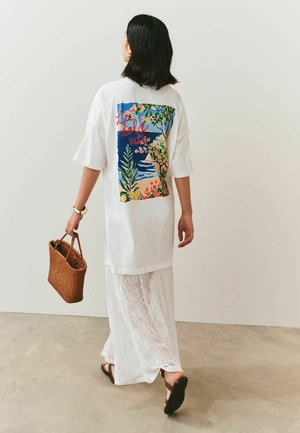 Woman walking away wearing white dress with colorful coastal print and text "La dolce vita," carrying brown woven handbag and wearing sandals.