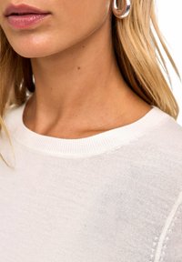 Jasnobeżowy sweter z okrągłym dekoltem, prążkowanym kołnierzem i gładką fakturą. Widok z bliska na ramię i linię szyi, podkreślający prostotę.