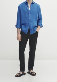 Man i en uppvikt blå knappskjorta, svarta byxor och svarta sandaler som står mot en enhetlig ljus bakgrund.