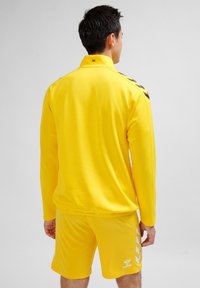 Gul sportsoverall som består av en långärmad tröja och shorts, tillverkad av ett mjukt tyg. Svarta accentlinjer på axlarna; logotyp synlig.