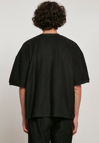 Tricou lung negru cu dungi, supradimensionat, cu mâneci scurte, prezentând o croială lejeră și o textură netedă, văzut din spate.