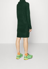 Abito verde lavorato a maglia con collo alto, abbinato a scarpe da ginnastica verde brillante e calzini arancioni. L'abito ha una vestibilità ampia e bordi a costine.
