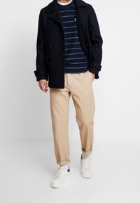 Homme portant un caban bleu marine, un pull rayé bleu marine, un pantalon beige retroussé aux ourlets, des chaussettes blanches et des baskets blanches avec des accents bleus marines.