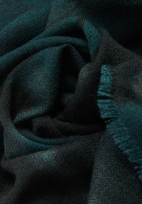 Tejido texturizado en verde y negro, colgado de manera suelta para mostrar un patrón tejido y bordes con flecos, creando una apariencia suave y acogedora.