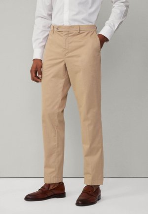 Pantaloni in cotone beige chiaro con taglio dritto, chiusura con bottone e tasche laterali, abbinati a scarpe in pelle marrone scuro.