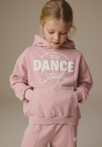 Rožinė gobtuvo palaidinė su balta užrašu „DANCE Studio“ ir kengūros kišene, pagaminta iš minkštos medžiagos. Raišteliai ant rankovių ir apykaklės. Suderinti kelnes.