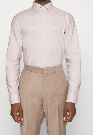 Mężczyzna ubrany w jasnoróżową, długorękawową koszulę zapinaną na guziki z logo „BOSS” oraz beżowe, dopasowane spodnie, stojący na jednolitym tle.