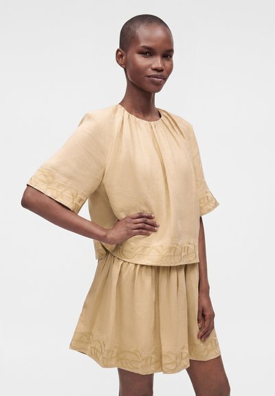 Femme aux cheveux courts portant une tenue beige deux-pièces avec des bords brodés, debout avec une main sur la hanche sur un fond uni.
