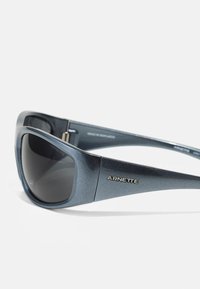 Occhiali da sole con lenti rettangolari di grandi dimensioni e tonalità blu, caratterizzati da una texture lucida e un logo "ARNETTE" in argento sulla montatura.