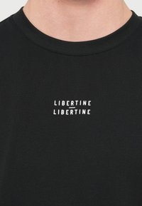 Μαύρο βαμβακερό μπλουζάκι με στρογγυλή λαιμόκοψη· φέρει λευκό κείμενο "LIBERTINE LIBERTINE" τυπωμένο στο στήθος σε οριζόντια διάταξη.